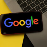 Telefon z włączonym logo Google. Leży na skarwku laptopa. Ten z kolei, na kolorowej podkładce.